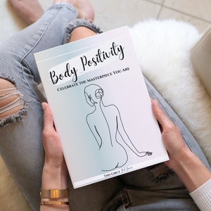 May include: A white journal with the text "Body Positivity Celebrate the Masterpiece You Are" and a line drawing of a person's back. The journal is being held by a person wearing jeans and a white sweater.