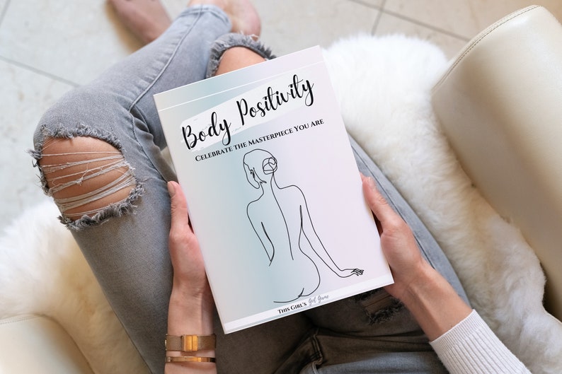 May include: A white journal with the text "Body Positivity Celebrate the Masterpiece You Are" and a line drawing of a person's back. The journal is being held by a person wearing jeans and a white sweater.