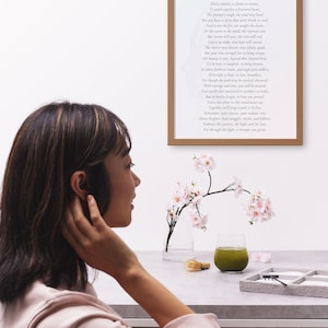 May include: A framed print with a poem in black text. A glass of green liquid, a small whisk, and a tray with sand and small rocks are on a table. Pink flowers in a clear vase are also on the table. A person is in the foreground.