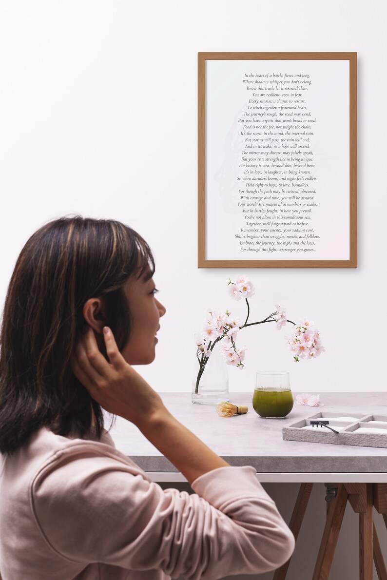 May include: A framed print with a poem in black text. A glass of green liquid, a small whisk, and a tray with sand and small rocks are on a table. Pink flowers in a clear vase are also on the table. A person is in the foreground.