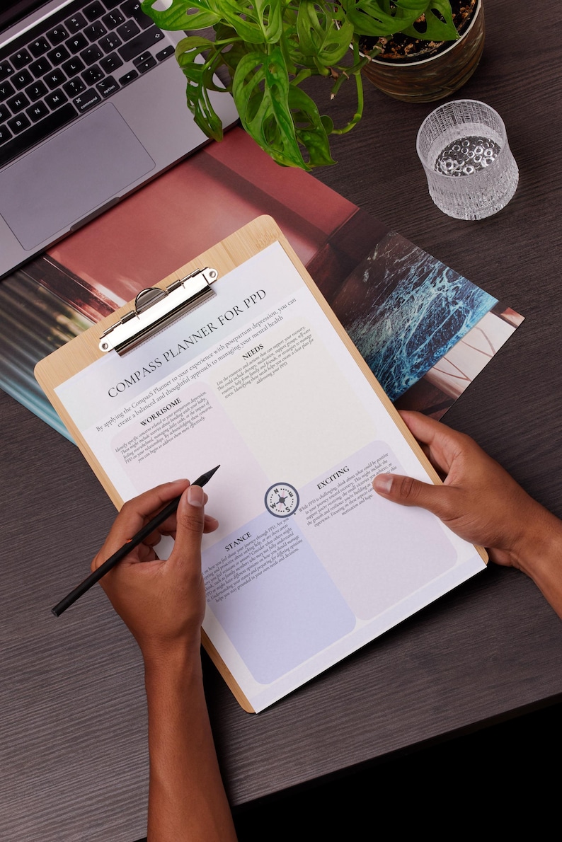 May include: A wooden clipboard with a printed planner titled "COMPASS PLANNER FOR PPD" is held by a person. The planner has sections labeled "Worrysome", "Needs", "Stance", and "Exciting". A black pen is poised over the paper.