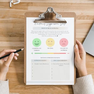 May include: A clipboard with an "Emotional Suppression Scale" chart, featuring smiley faces and a sad face, is held by a person. A black pencil rests in a hand, and a laptop and notebook are also on the wooden surface.