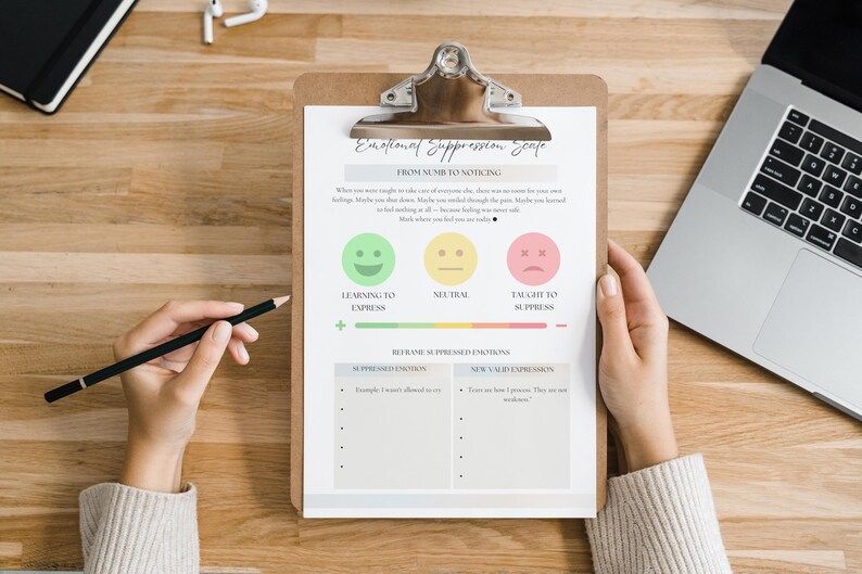 May include: A clipboard with an "Emotional Suppression Scale" chart, featuring smiley faces and a sad face, is held by a person. A black pencil rests in a hand, and a laptop and notebook are also on the wooden surface.