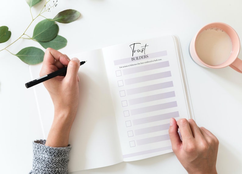 Puede incluir: Una libreta blanca con una lista de verificaci&oacute;n titulada "Trust Builders" con el texto "List actions or behaviors that have strengthened or built trust." Una persona est&aacute; escribiendo en la libreta con un bol&iacute;grafo negro.
