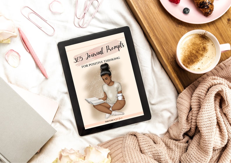 Puede incluir: Una tableta negra con una cubierta rosa y blanca que muestra el texto "365 Journal Prompts for Positive Thinking". La cubierta presenta una ilustraci&oacute;n de una mujer leyendo un libro y sosteniendo una taza de caf&eacute;.