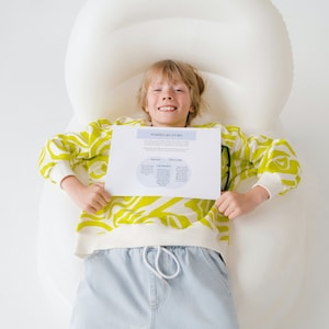May include: A young person wearing a yellow and white patterned sweater and blue jeans is lying on a white inflatable lounger. They are holding a white sheet of paper with the text "Personal and Family Stories" and a diagram with circles and text.