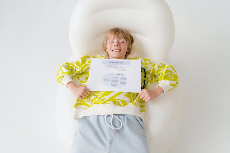 May include: A young person wearing a yellow and white patterned sweater and blue jeans is lying on a white inflatable lounger. They are holding a white sheet of paper with the text "Personal and Family Stories" and a diagram with circles and text.