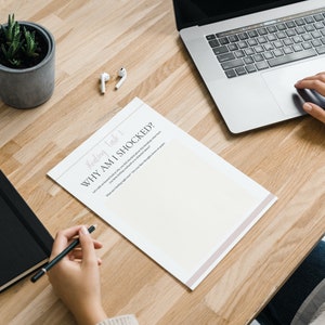 May include: A person is sitting at a wooden desk with a laptop, a notebook, a potted plant, and a sheet of paper with the title "Weekly Task 1: Why Am I Shocked?" The person is writing on the paper with a pen.