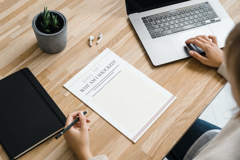May include: A person is sitting at a wooden desk with a laptop, a notebook, a potted plant, and a sheet of paper with the title "Weekly Task 1: Why Am I Shocked?" The person is writing on the paper with a pen.