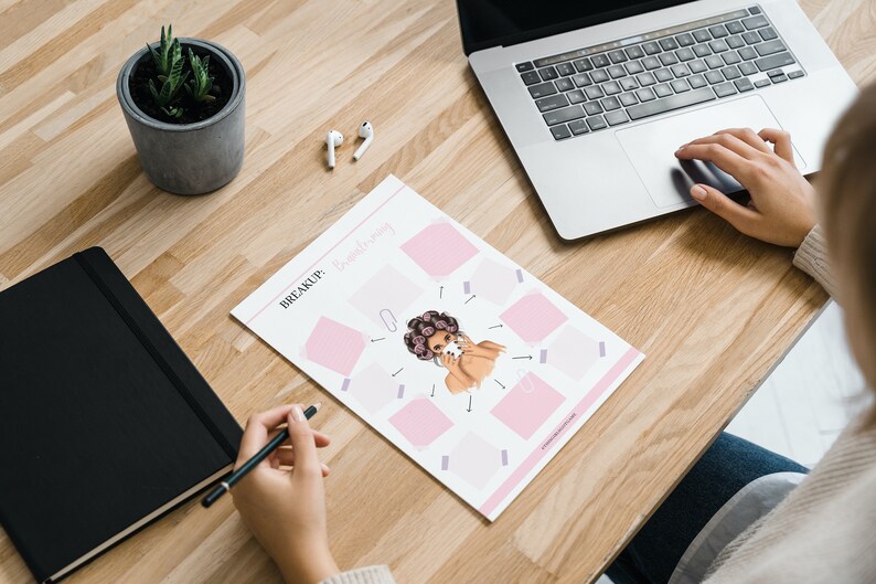 May include: A printable worksheet with the text "BREAKUP: Brainstorming" and a cartoon illustration of a woman with pink hair and a pink background. The worksheet is on a wooden table with a laptop, a potted plant, and a pair of wireless earbuds.