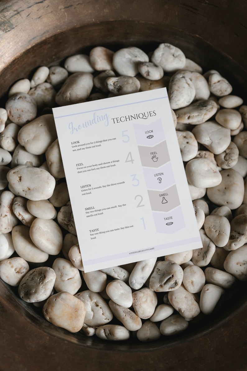 May include: A white card with the title "Grounding Techniques" and a list of five grounding techniques. The techniques are numbered 1 through 5 and include: Taste, Smell, Listen, Feel, and Look. Each technique has a corresponding icon next to it. The card is lying on a bed of white rocks.