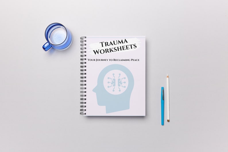 May include: A spiral-bound notebook with the title "TRAUMA WORKSHEETS" and the tagline "YOUR JOURNEY TO RECLAIMING PEACE." The cover features a light blue silhouette of a head with a brain-like design. A blue glass cup, a pen, and a pencil are also on the white surface.