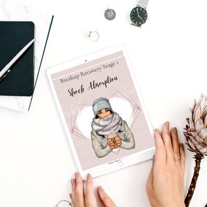 May include: A digital tablet displaying a pink and white graphic with the text "Breakup Recovery Stage 1: Shock Absorption". The graphic features a woman wearing a gray hat, scarf, and jacket, holding a cup of coffee.