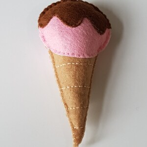 May include: A handmade felt ice cream cone with pink ice cream and chocolate fudge topping. The cone is tan and stitched with light brown thread. The ice cream is a soft pink color.