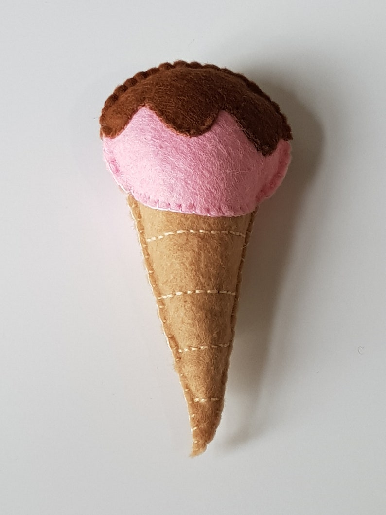 May include: A handmade felt ice cream cone with pink ice cream and chocolate fudge topping. The cone is tan and stitched with light brown thread. The ice cream is a soft pink color.