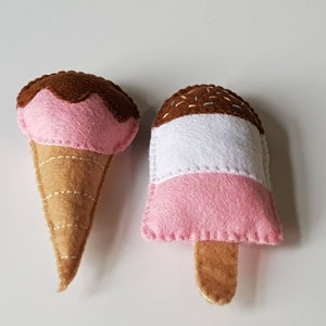 May include: Two felt ice cream treats: a cone with pink ice cream and chocolate topping, and a popsicle with pink, white, and chocolate layers. Both are handmade with stitched details, perfect for a child's play food collection.