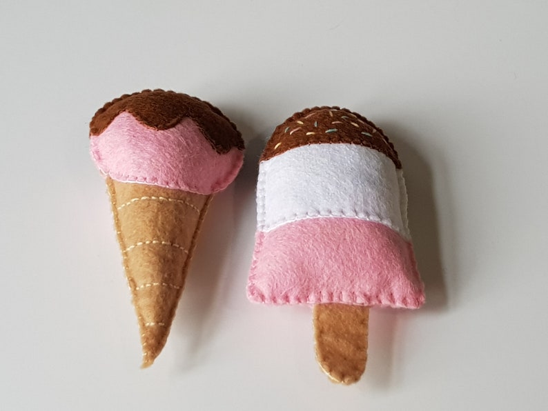 May include: Two felt ice cream treats: a cone with pink ice cream and chocolate topping, and a popsicle with pink, white, and chocolate layers. Both are handmade with stitched details, perfect for a child's play food collection.