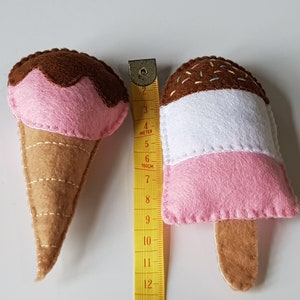 May include: Two felt ice cream toys. One is a cone with a pink scoop topped with brown felt. The other is a popsicle with pink, white, and brown layers. A yellow measuring tape is next to the toys.