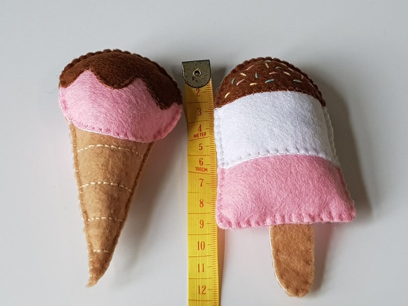 May include: Two felt ice cream toys. One is a cone with a pink scoop topped with brown felt. The other is a popsicle with pink, white, and brown layers. A yellow measuring tape is next to the toys.