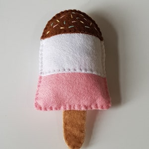 May include: A handmade felt ice pop in pink, white, and brown, with a wooden stick. The top section is brown with colorful sprinkles, the middle is white, and the bottom is pink. The edges are stitched with white thread.