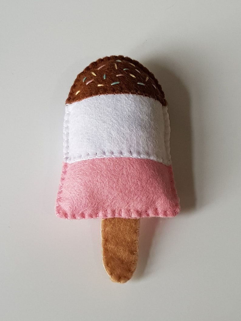 May include: A handmade felt ice pop in pink, white, and brown, with a wooden stick. The top section is brown with colorful sprinkles, the middle is white, and the bottom is pink. The edges are stitched with white thread.