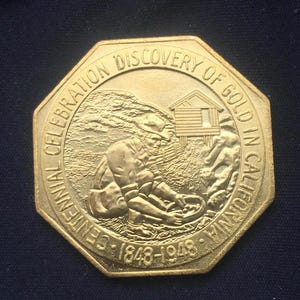 May include: Gold-colored commemorative coin celebrating the centennial of the discovery of gold in California. The coin features a depiction of a gold miner panning for gold, a small cabin, and the text "Centennial Celebration Discovery of Gold in California 1848-1948".