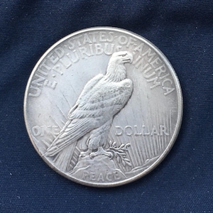 May include: A silver Peace Dollar coin with the inscription "United States of America E Pluribus Unum One Dollar Peace" and a bald eagle with outstretched wings.