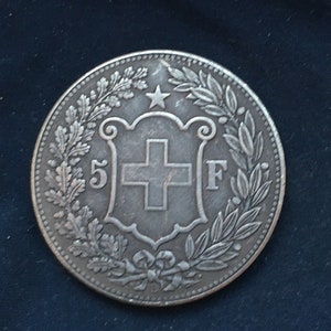 Op de afbeelding: Een zilveren munt met een krans van bladeren rond een schild met een kruis en het getal "5" en de letter "F".