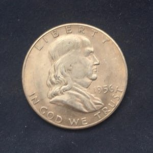 Könnte beinhalten: Eine bronzefarbene Münze mit einem Profil von Benjamin Franklin, den Inschriften "LIBERTY", "IN GOD WE TRUST" und dem Jahr "1956A". Die Münze ist kreisförmig und weist leichte Abnutzungserscheinungen auf.