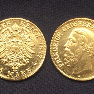 May include: Two gold coins. One coin features an eagle emblem and the text "DEUTSCHES REICH 1877 5 MARK". The other coin shows a profile of a man and the text "FRIEDRICH GROSHERZOG VON BADEN". Both coins are on a dark blue surface.