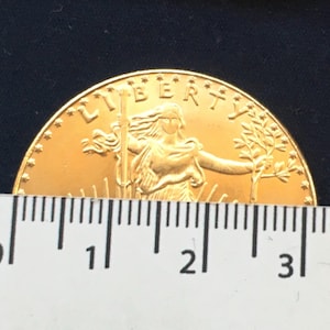 May include: A gold-colored coin with the word "LIBERTY" inscribed around the edge. The coin features a woman holding a torch and a branch, with rays of light emanating from behind her. The coin is measured against a ruler, showing it is approximately 3 inches in diameter.