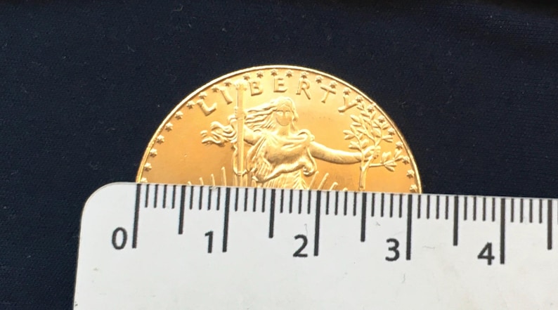 May include: A gold-colored coin with the word "LIBERTY" inscribed around the edge. The coin features a woman holding a torch and a branch, with rays of light emanating from behind her. The coin is measured against a ruler, showing it is approximately 3 inches in diameter.