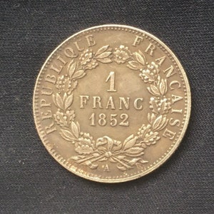 May include: A silver French 1 Franc coin from 1852. The coin features a wreath of leaves and the words "Republique Francaise" around the edge.