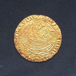 Könnte beinhalten: Eine Goldmünze mit einem detaillierten Design eines Schiffes und einer Krone. Die Münze hat einen erhöhten Rand und eine glatte Oberfläche.