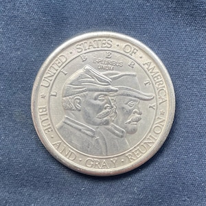 Pode incluir: Uma moeda comemorativa de tom dourado com os perfis de dois soldados de chapéu. O texto diz UNITED STATES OF AMERICA, LIBERTY, E PLURIBUS UNUM e BLUE AND GRAY REUNION. A moeda tem uma borda circular e um acabamento metálico.