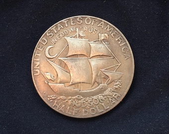 May include: A bronze Hudson Half Dollar coin featuring a sailing ship with multiple sails. The coin is inscribed with "United States of America" and "In God We Trust". The coin is set against a dark background.