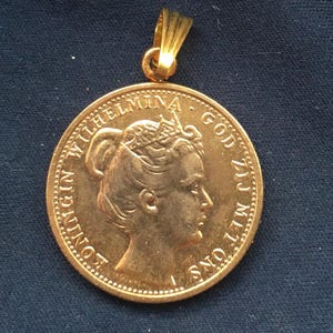 May include: A gold-colored pendant featuring a detailed profile of a woman and the inscription "KONINGIN WILHELMINA GOD ZIJ MET ONS." The pendant has a loop for a chain and is set against a dark blue background.