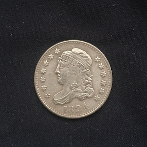 May include: A silver coin from 1824, featuring a profile of a woman wearing a cap with the word "LIBERTY" on it. The coin is surrounded by small star-shaped designs. The coin is on a black background.