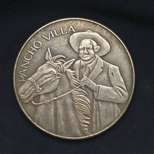 May include: Silver coin featuring a portrait of Pancho Villa, a Mexican revolutionary general, on horseback. The coin has the words "Pancho Villa" inscribed around the edge.