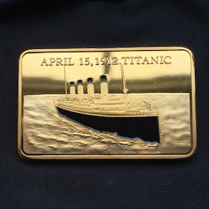 May include: A rectangular gold-colored metal plaque featuring a relief of the Titanic ship on water. The plaque has the text "APRIL 15, 1912. TITANIC" at the top. The ship is black and white with four smokestacks.