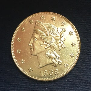 Peut inclure: Une pièce de monnaie dorée avec le profil d'une femme portant un couvre-chef et l'année 1868. La pièce a un bord détaillé et est ornée d'étoiles. Le mot "LIBERTY" est visible sur le couvre-chef.
