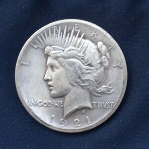 May include: A silver Peace dollar coin with a portrait of Liberty on the obverse. The coin is dated 1921 and has the inscription "IN GOD WE TRUST" and "LIBERTY" on the obverse. The coin is in good condition.