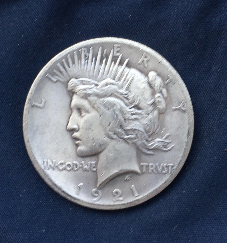 May include: A silver Peace dollar coin with a portrait of Liberty on the obverse. The coin is dated 1921 and has the inscription "IN GOD WE TRUST" and "LIBERTY" on the obverse. The coin is in good condition.
