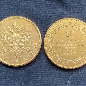 May include: Two gold Finnish coins. One coin features a detailed coat of arms with a double-headed eagle and the word FINLAND. The other coin has the text "20 MARKKA 1915" and other inscriptions. The coins are on a dark blue surface.