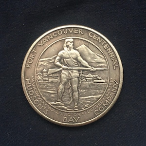 May include: A bronze-colored commemorative coin with the text "Fort Vancouver Centennial" and "Hudson's Bay Company". The coin features an image of a man holding a rifle, with a fort and mountain in the background. The coin is dated 1825.