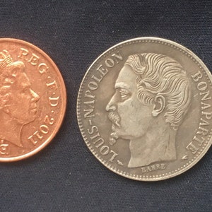May include: Two coins, one copper and one silver, with portraits of Queen Elizabeth II and Louis Napoleon Bonaparte. The copper coin has the inscription "ELIZABETH II D.G. REG. F.D. 2011" and the silver coin has the inscription "LOUIS-NAPOLEON BONAPARTE EMPEREUR".
