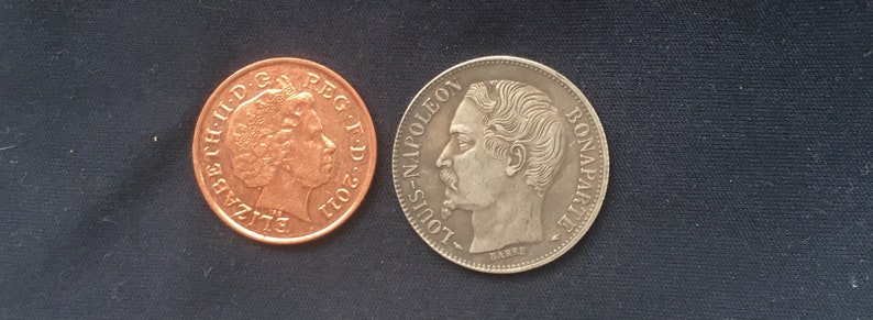 May include: Two coins, one copper and one silver, with portraits of Queen Elizabeth II and Louis Napoleon Bonaparte. The copper coin has the inscription "ELIZABETH II D.G. REG. F.D. 2011" and the silver coin has the inscription "LOUIS-NAPOLEON BONAPARTE EMPEREUR".