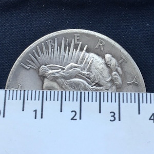 May include: A silver Peace Dollar coin with the image of Liberty on the obverse side. The coin is being measured with a ruler, showing it is approximately 3 inches in diameter.