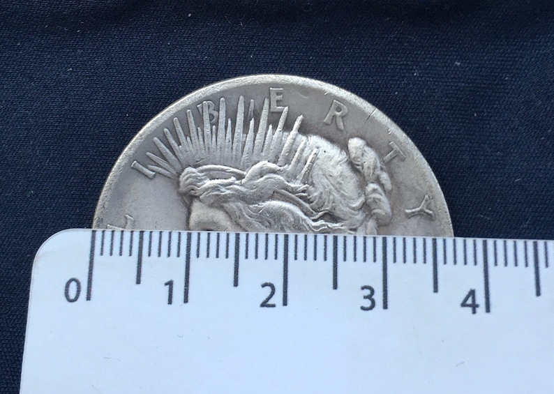 May include: A silver Peace Dollar coin with the image of Liberty on the obverse side. The coin is being measured with a ruler, showing it is approximately 3 inches in diameter.