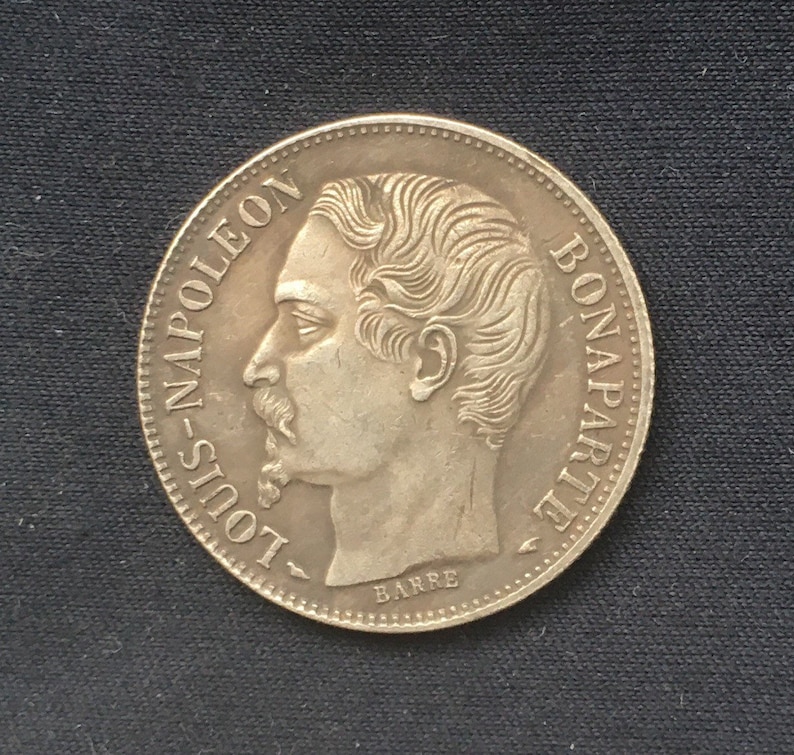 May include: Silver coin featuring a profile portrait of Louis Napoleon Bonaparte, with the inscription "LOUIS-NAPOLEON BONAPARTE" around the edge and "BARRE" at the bottom.